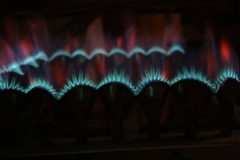 A technician's gloved hand holding a new silicon nitride igniter next to a gas furnace, ready to replace a furnace igniter
